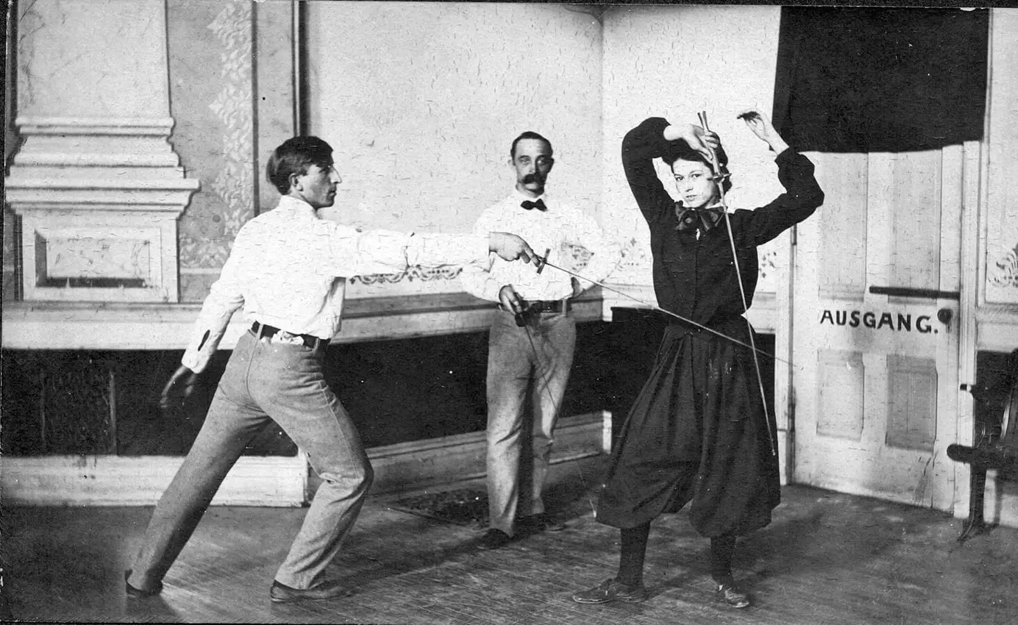 Photo of a fencing instructor looking on as a man advances on a woman parrying her sword while looking at the camera.