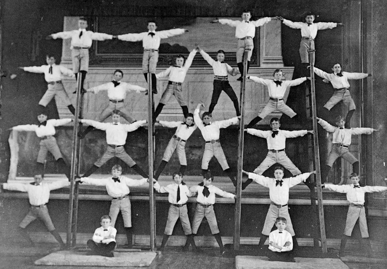 24 boys are shown, balancing on top of one another and on ladders. They form groups of three on either side of the ladders, and each of the four ladders has one boy perched on top. Two boys sit on the ground in front of the scene.