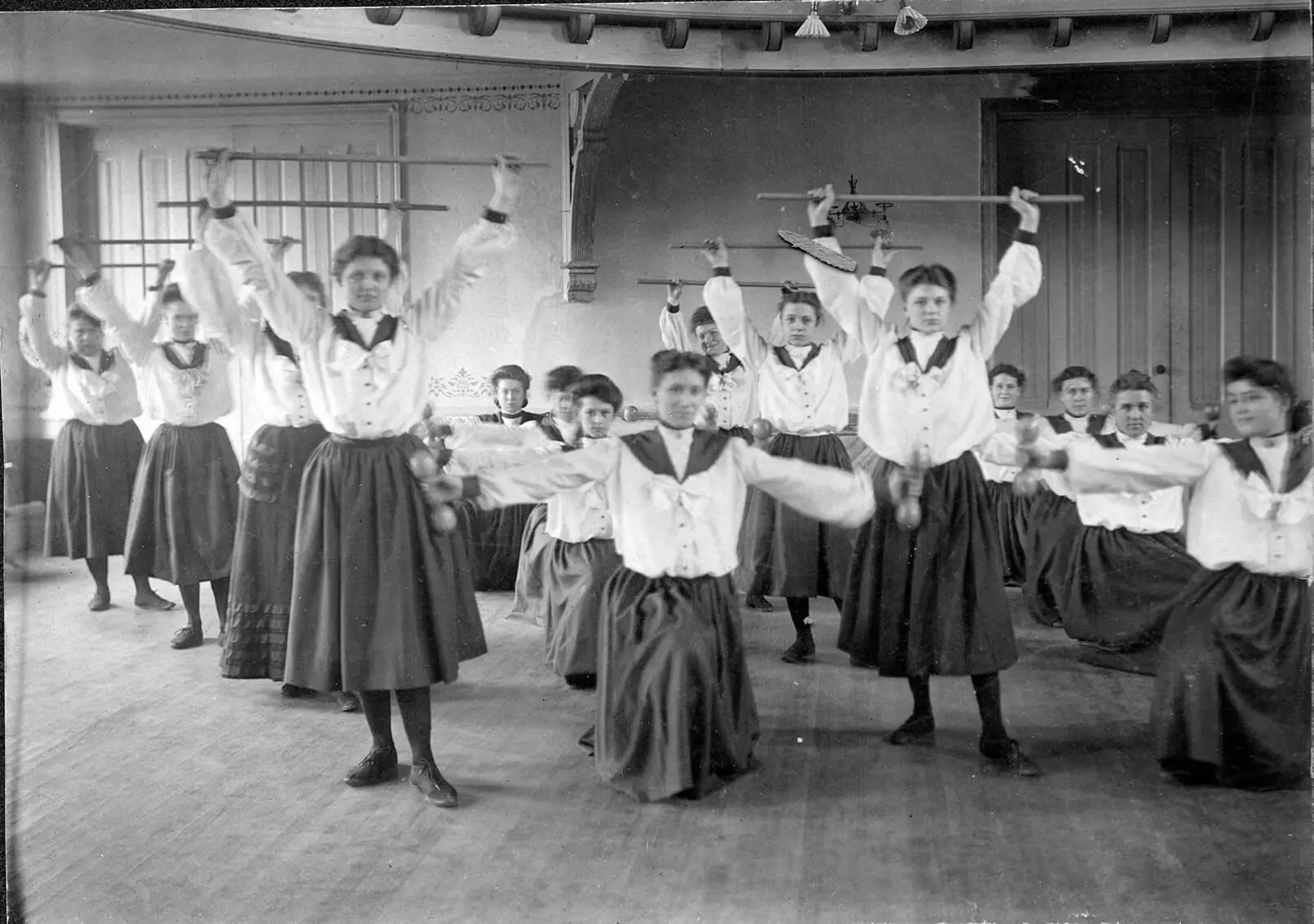Black and white photo of women standing in four rows. From left to right, rows one and three stretch upwards with a rod in hand and rows two and four lunge with dumbbells held.