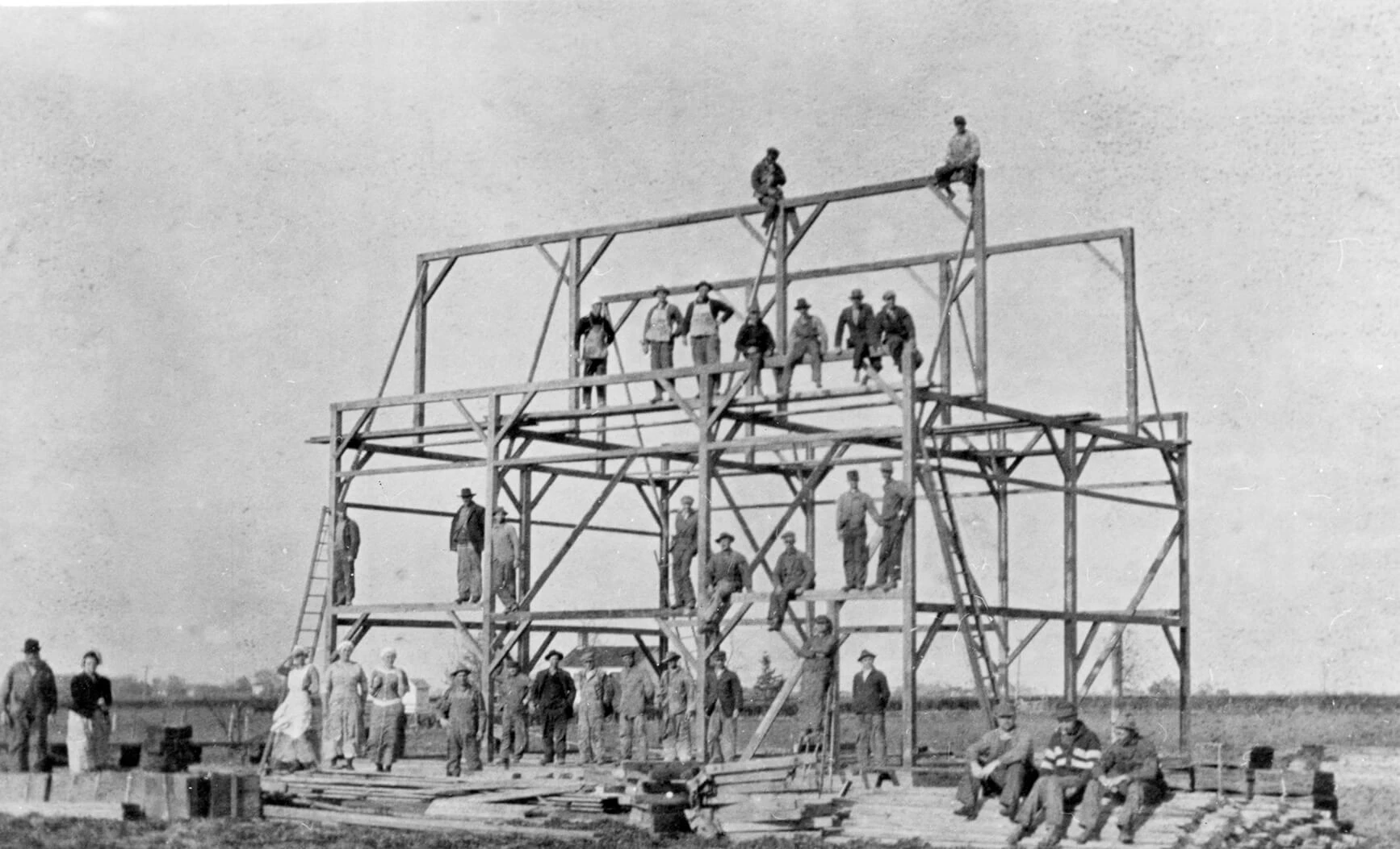 Photograph of a 3-story frame with men standing and sitting all over the frame, and on the ground. Two men are even on the very top of the roof beam... They must have confidence in their construction, that's for sure! There are approx. 25 men in the photo.