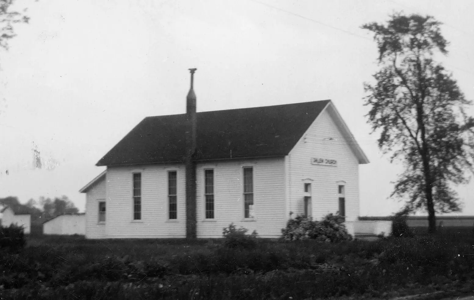 Black and white photo of a single-room wooden church, painted white, with two doors at the front.