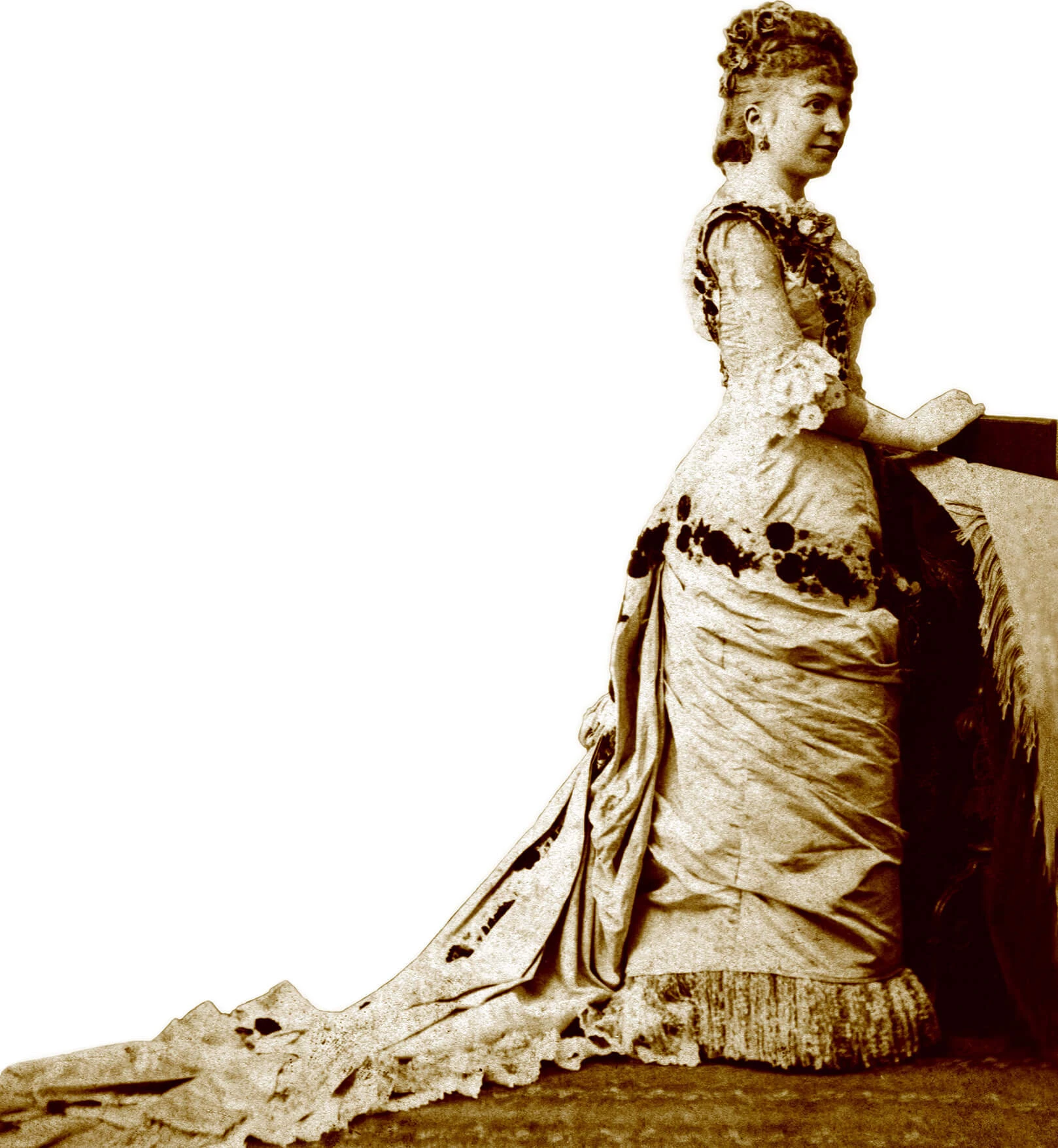 Full length portrait, showing her full gown and train. At the bottom of her skirt there are a few inches of fringe. The dress is light colored with darker floral details at the waist and shoulders. Sleeves go down to the elbow, where they end with two layers of lace flowing around the arm.