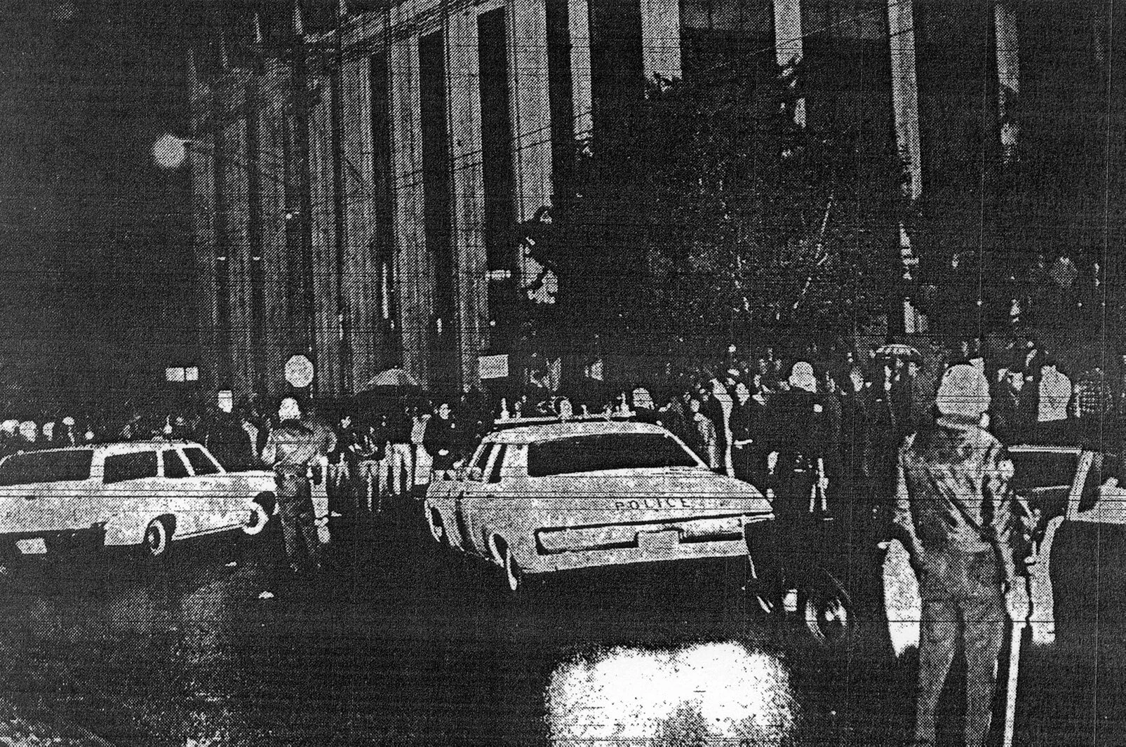 A police car and police man are seen in the foreground, and in the background are dozens of students. It is night time.