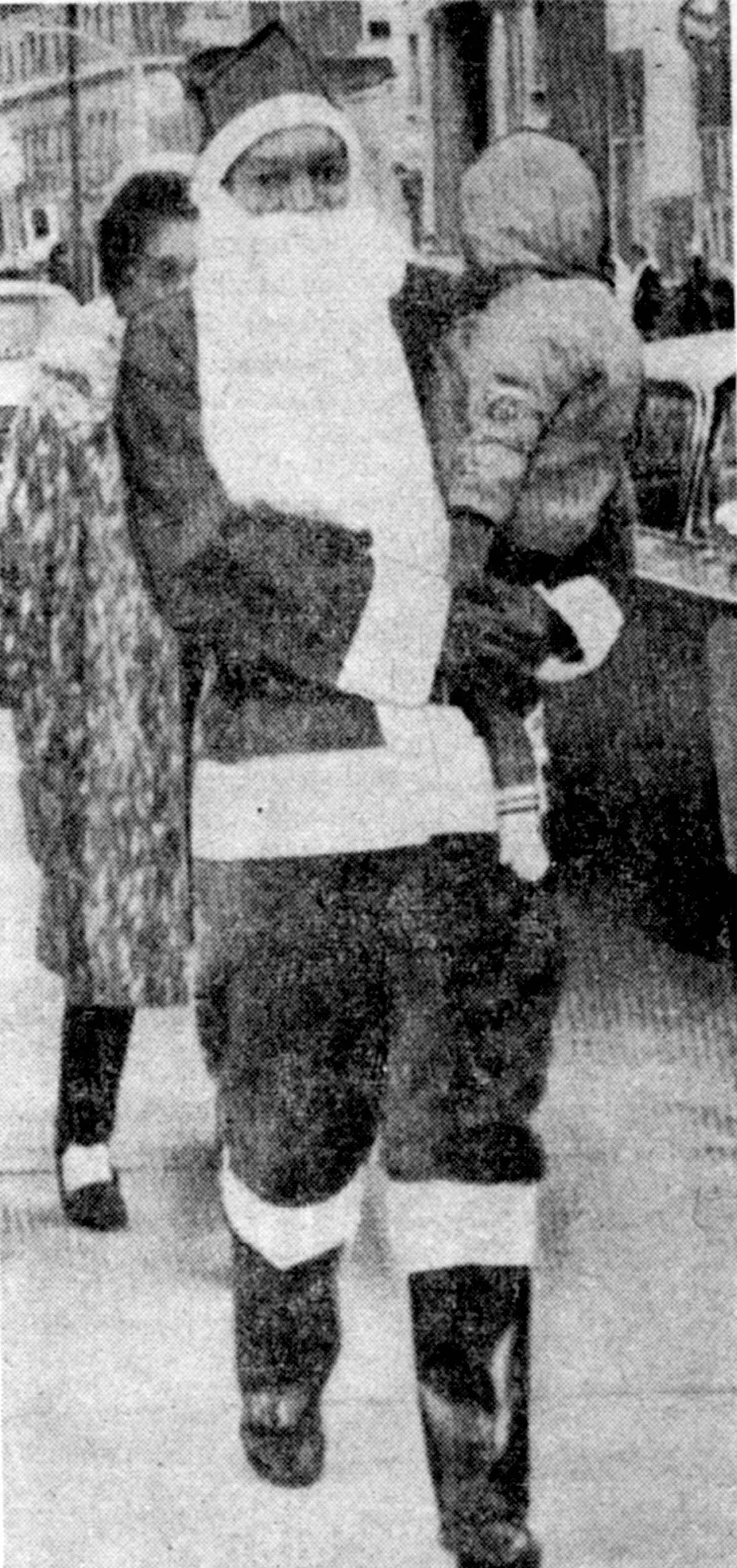 Merlin Kennedy is pictured wearing a Santa Claus suit, walking alongside the parade route while carrying a small child.