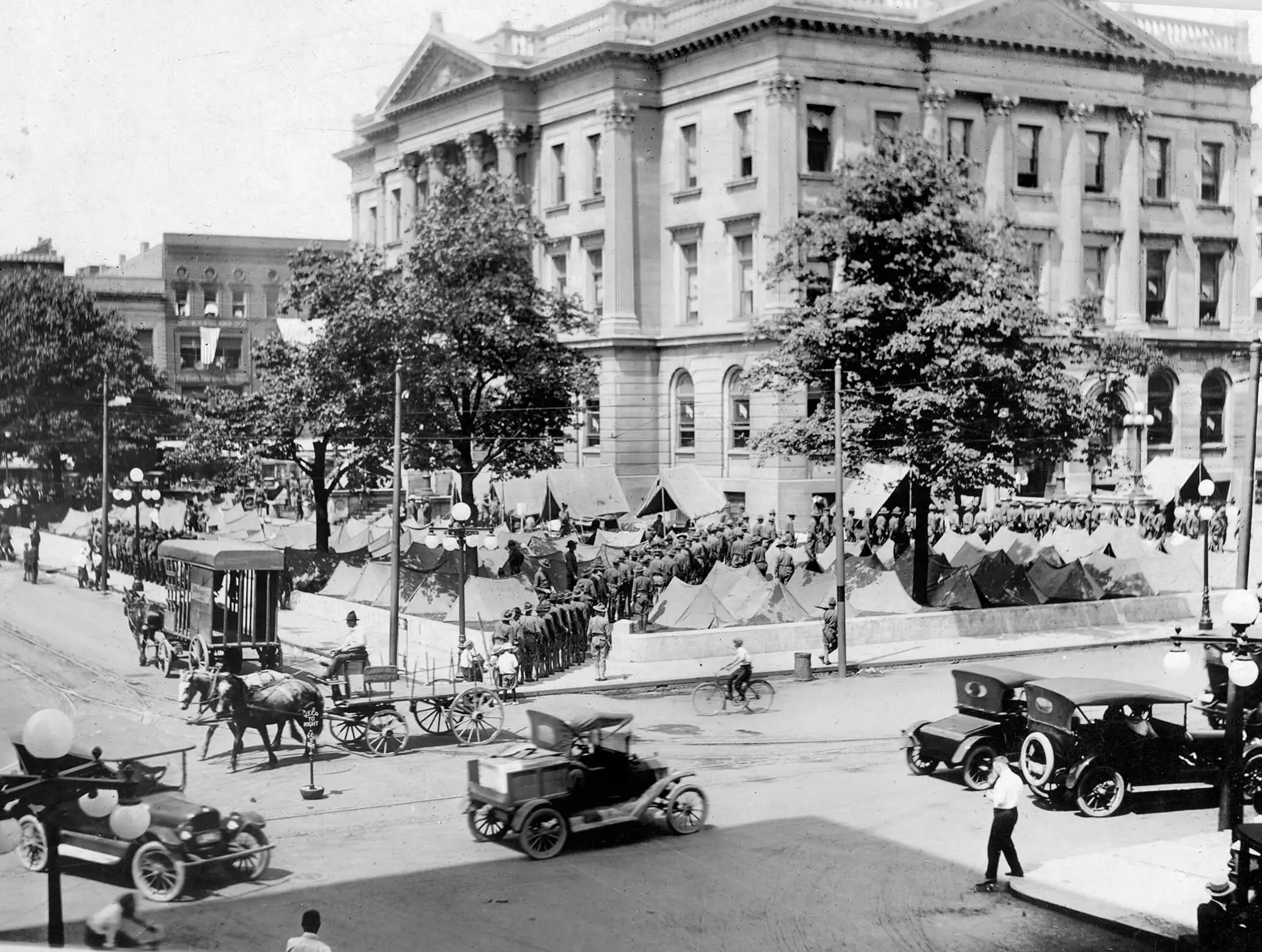 A three-story courthouse building with tents on the lawn out front. A long line of men in uniform extends from the building