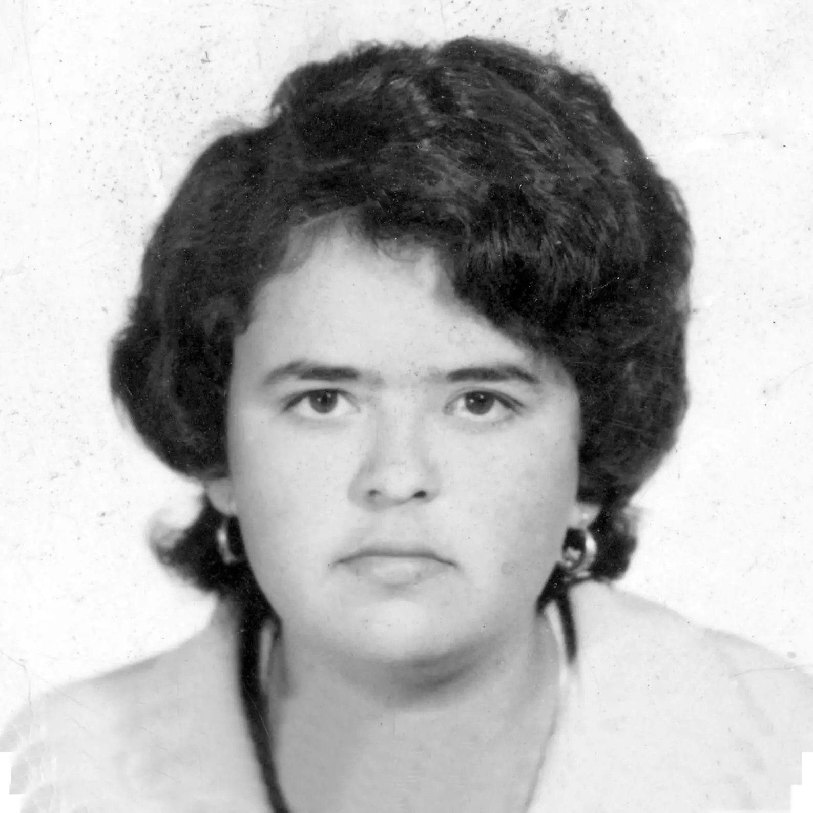 Black and white photo of a young woman with dark hair and dark eyes wearing hoop earrings. She is looking into the camera with a serious face, as instructed for passport photos.
