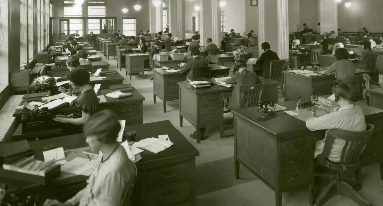 a large, open floorplan room with desks as far as the eye can see in all directions. At each desk a woman is either writing or typing at a typewriter. Most have short hair. The room has lots of natural light, and light figures also hang down from the ceiling.