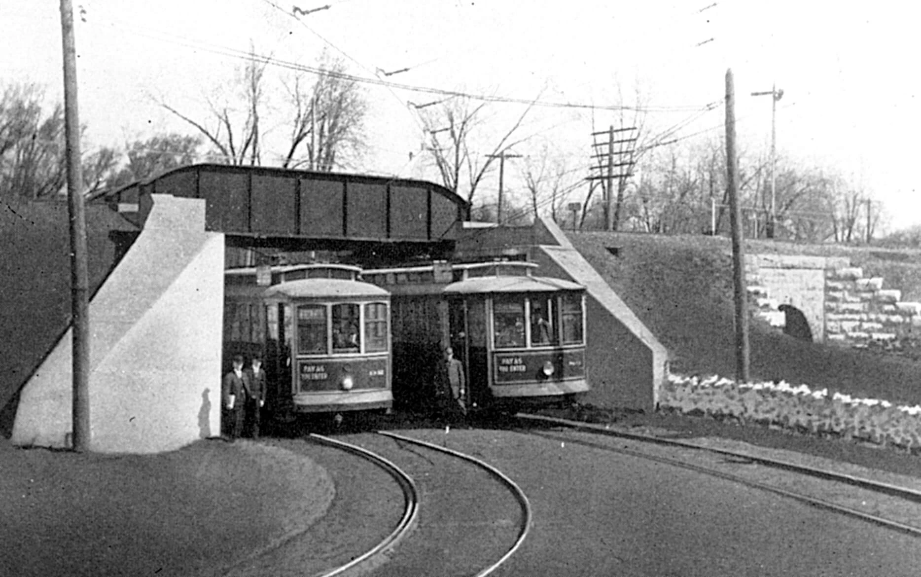 Black and white photo of two streetcars under a railroad overpass with three men dressed in conductor uniform standing off to the side