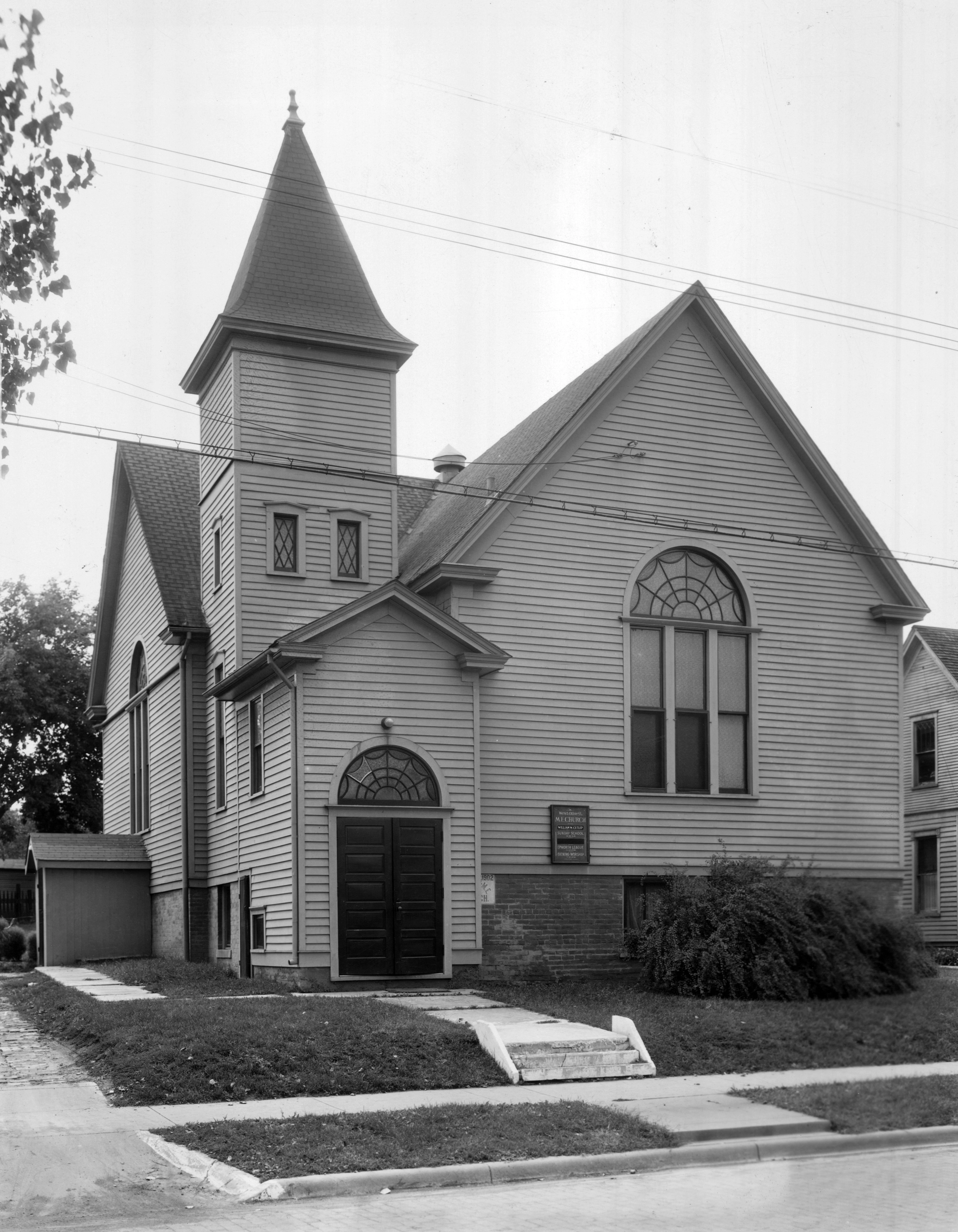 Black and white photo of a light colored church with wood siding and tall steeple.