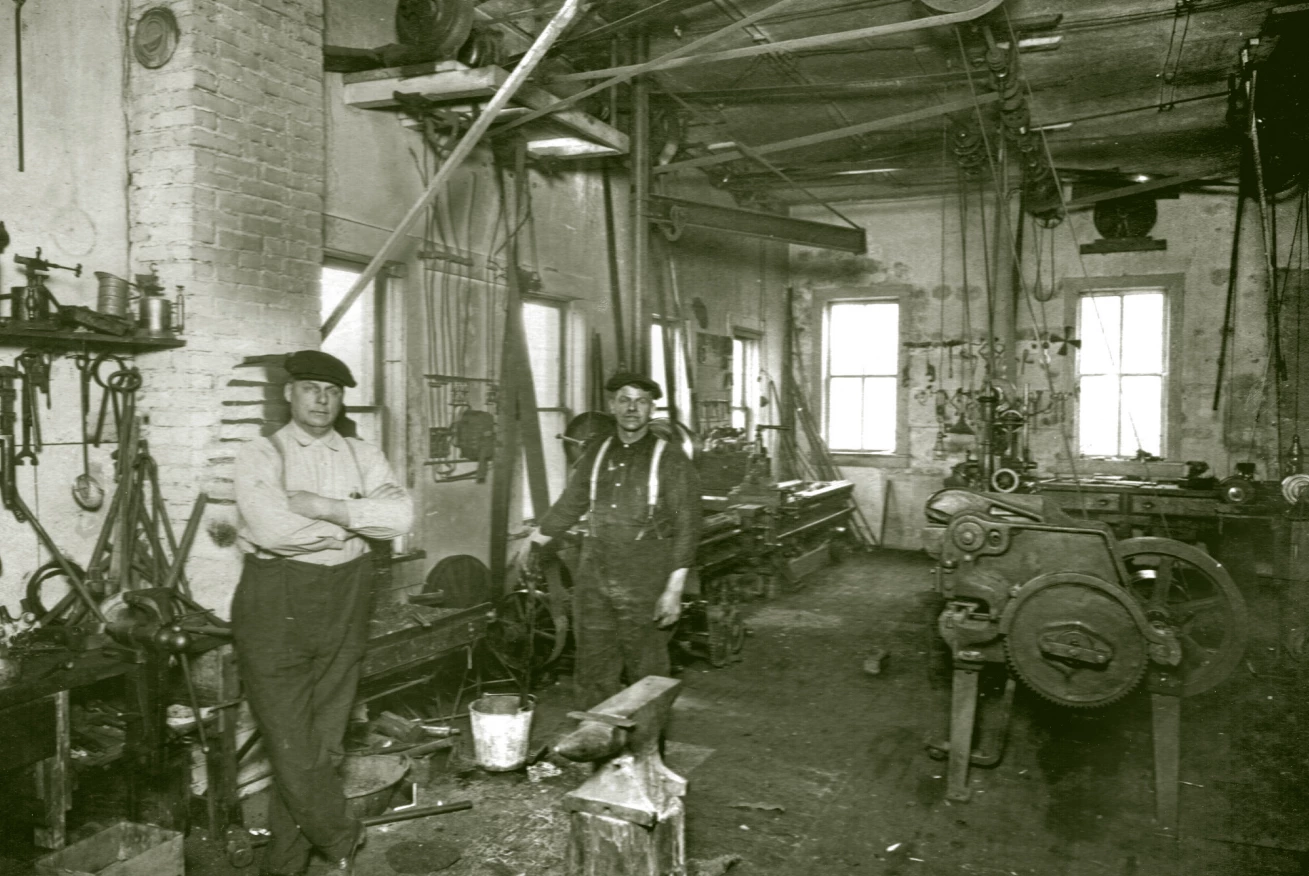 Two men stand inside a shop that has equipment and belts running all over. an anvil is in the foreground. There are windows and a high ceiling.