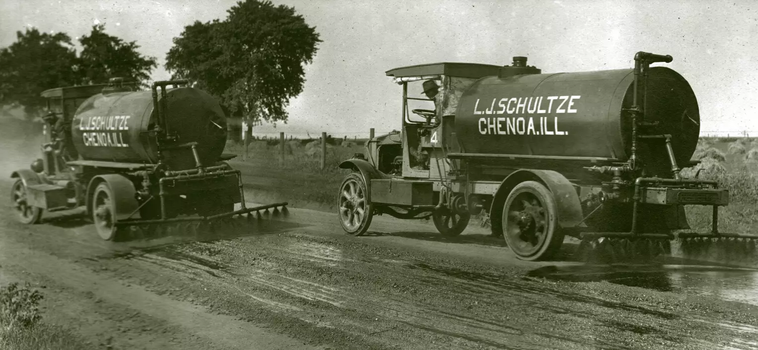 Two tank trucks drive down a rural road. Behind them are sprayers a foot or two off the ground. A black liquid is being sprayed out of the sprayers directly down.