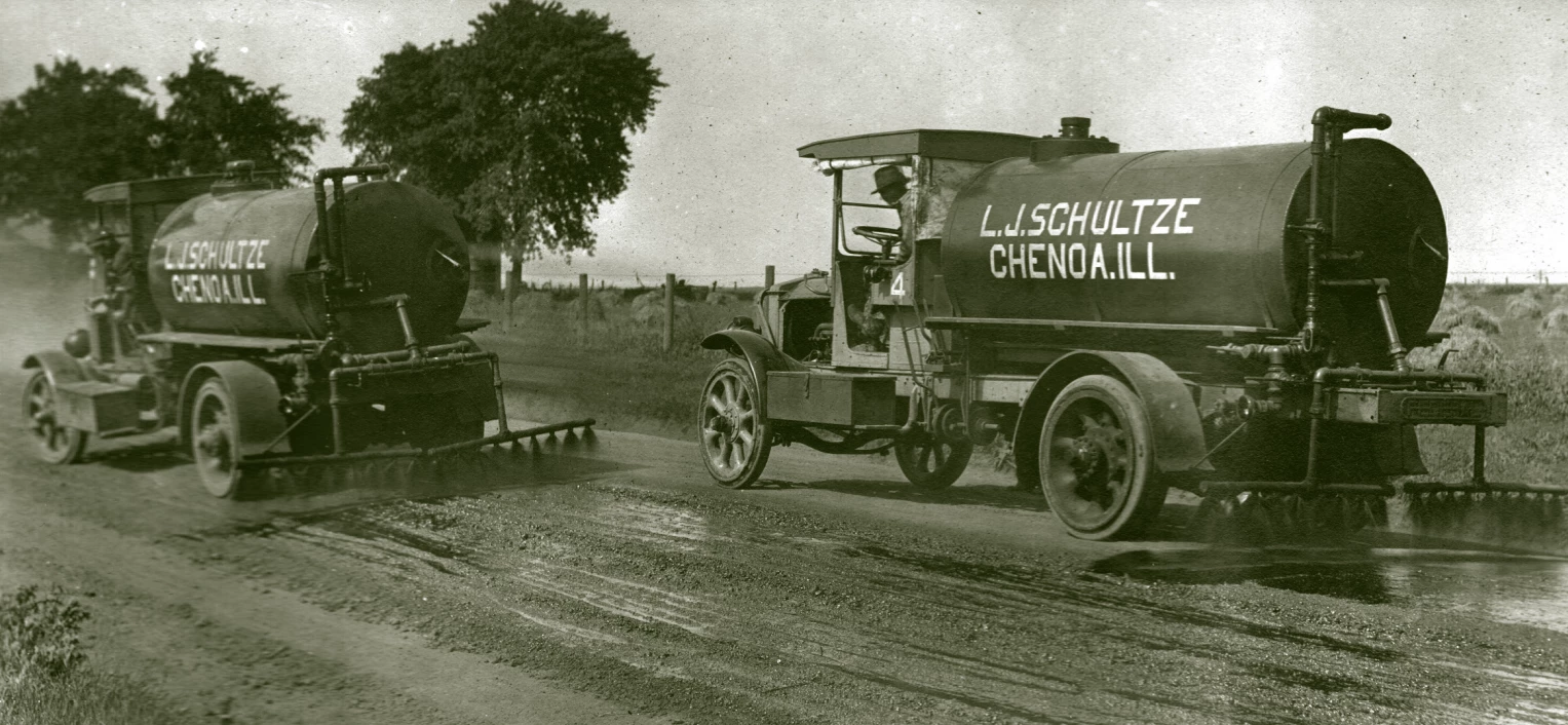 Two tank trucks drive down a rural road. Behind them are sprayers a foot or two off the ground. A black liquid is being sprayed out of the sprayers directly down.