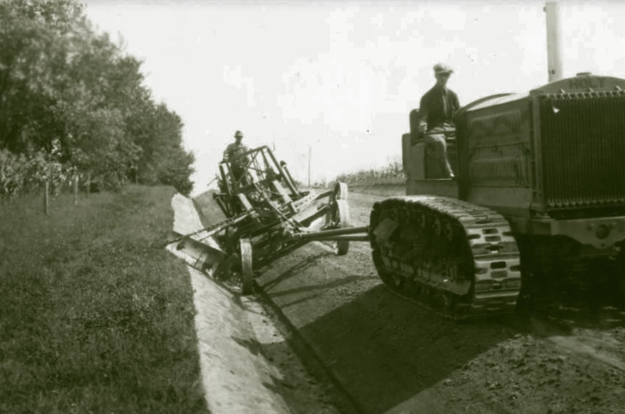 A large tractor with continuous track (not wheels) pulls a grader with a person on it. It appears they are on a rural road, there are large trees on one side and crops in the distance on the other side.