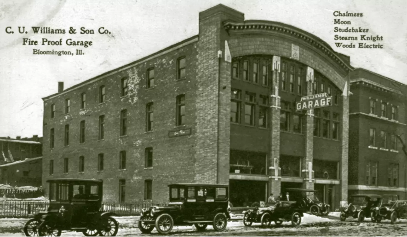 A 4-story brick building, along the top of the façade is a gentle arch spanning the width of the building. The first floor has three large garage doors, two of which appear to be open. There is snow on the ground and there are 6 cars lined up in front of the building.