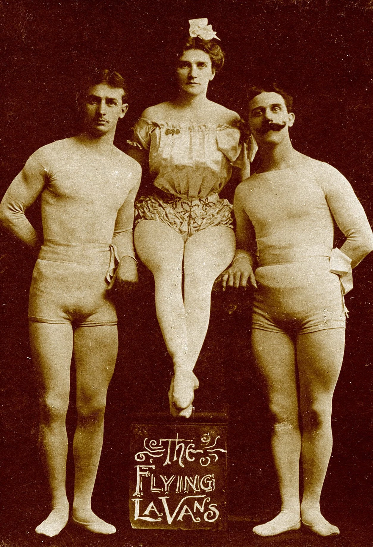 Full-length black and white photograph. The brothers are standing on either side and Amy is seated on something tall in the center. She is wearing a light colored off-the-shoulder ruffly shirt and ruffly short shorts, with a bead or sparkle detail around the hips. Tights cover the entire length of her legs. The men are wearing long-sleeved very tight lightly colored shirts and very short tight shorts. They are also wearing tights that cover the entire length of their legs and feet.