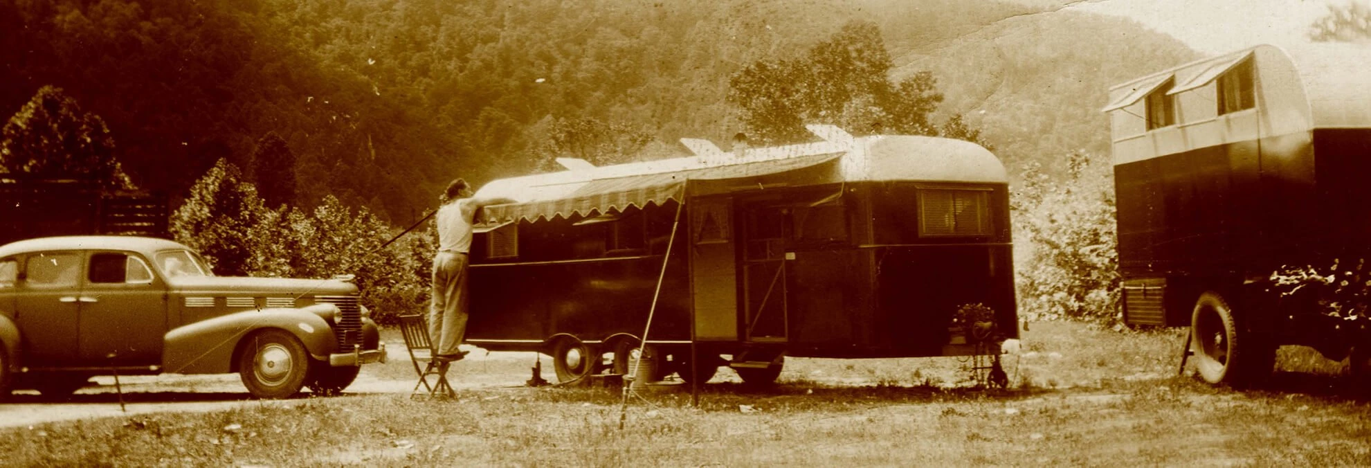 Two sleeping trailers are parked in the valley, mountains can be seen in the background, blocking the sky. A man stands on a wooden folding chair to attach or fix an awning attached to one of the trailers. A car is parked on the left.
