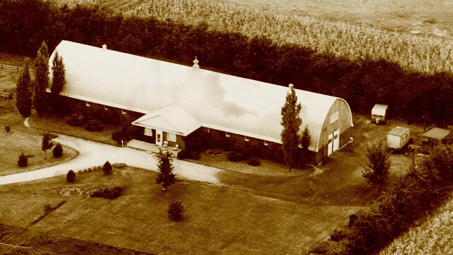A very long building with a curved roof. Entrance is right int he middle of the long side of the building, a sidewalk leads up to the door. Around the building are trees, bushes, and corn fields.