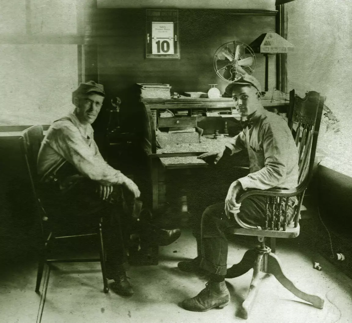 Two men seated in wooden chairs are a desk. They are looking at the camera. The desk has a fan, lamp, and some drawers.