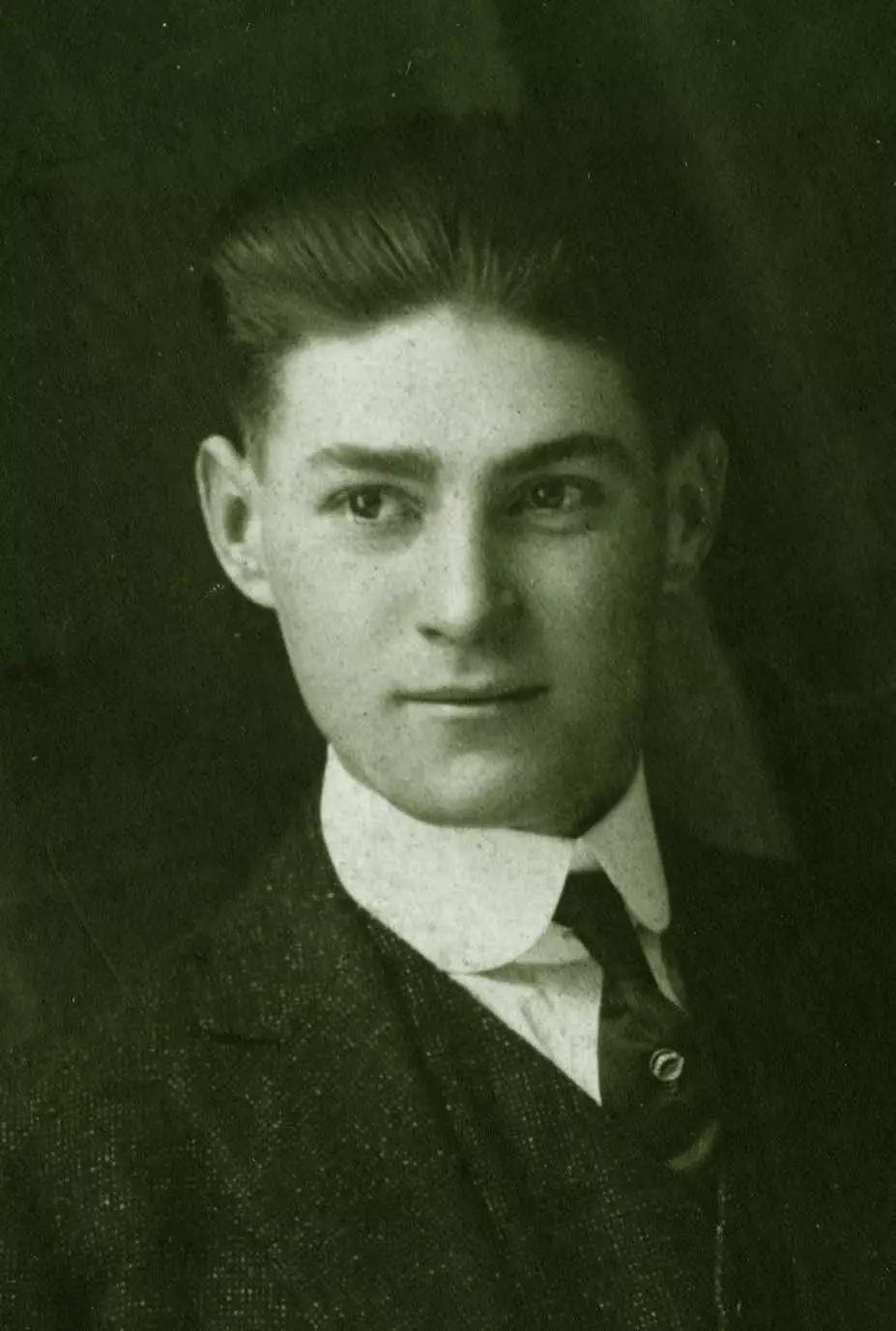 A young man in a suit and tie looks to the left of the camera. He has thick dark hair that is combed backwards.