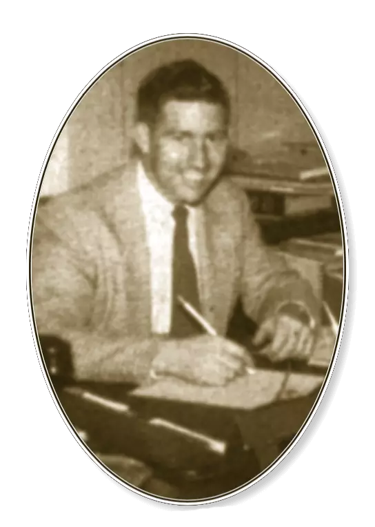 Grainy black and white photo of a light-skinned man in a light suit jacket and dark tie. He is sitting at a desk holding a pencil to paper. He is smiling and looking at the camera.