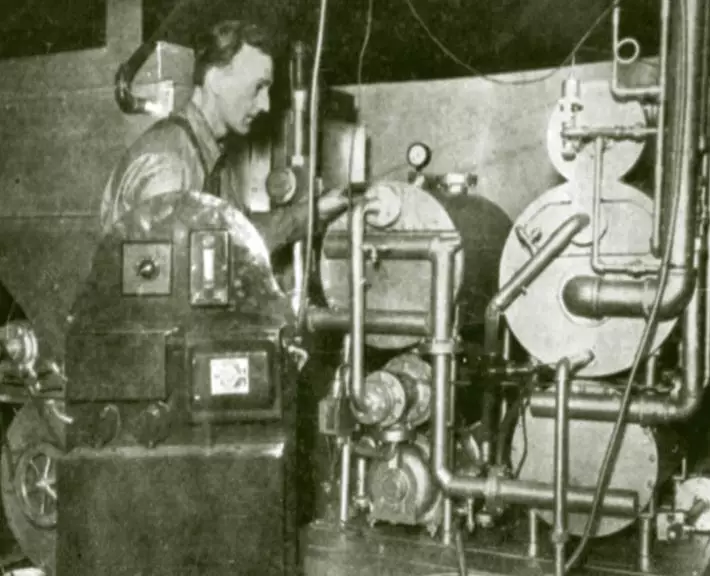 a man stands between tanks, which are interconnected by pipes. There are knobs, dials, and indicators on the tanks.