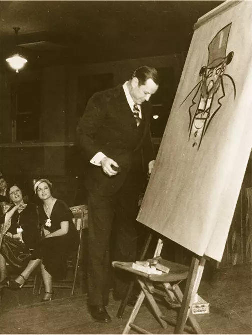 A light-skinned man in a dark suit and dark slicked back hair looks down as he stands in front of an easel with paper. On the paper he has drawn a rough sketch of an older man wearing a large tophat and suit. He appears to be drawing for a crowd, we can see two women looking on, they are seated at tables and wearing dresses.