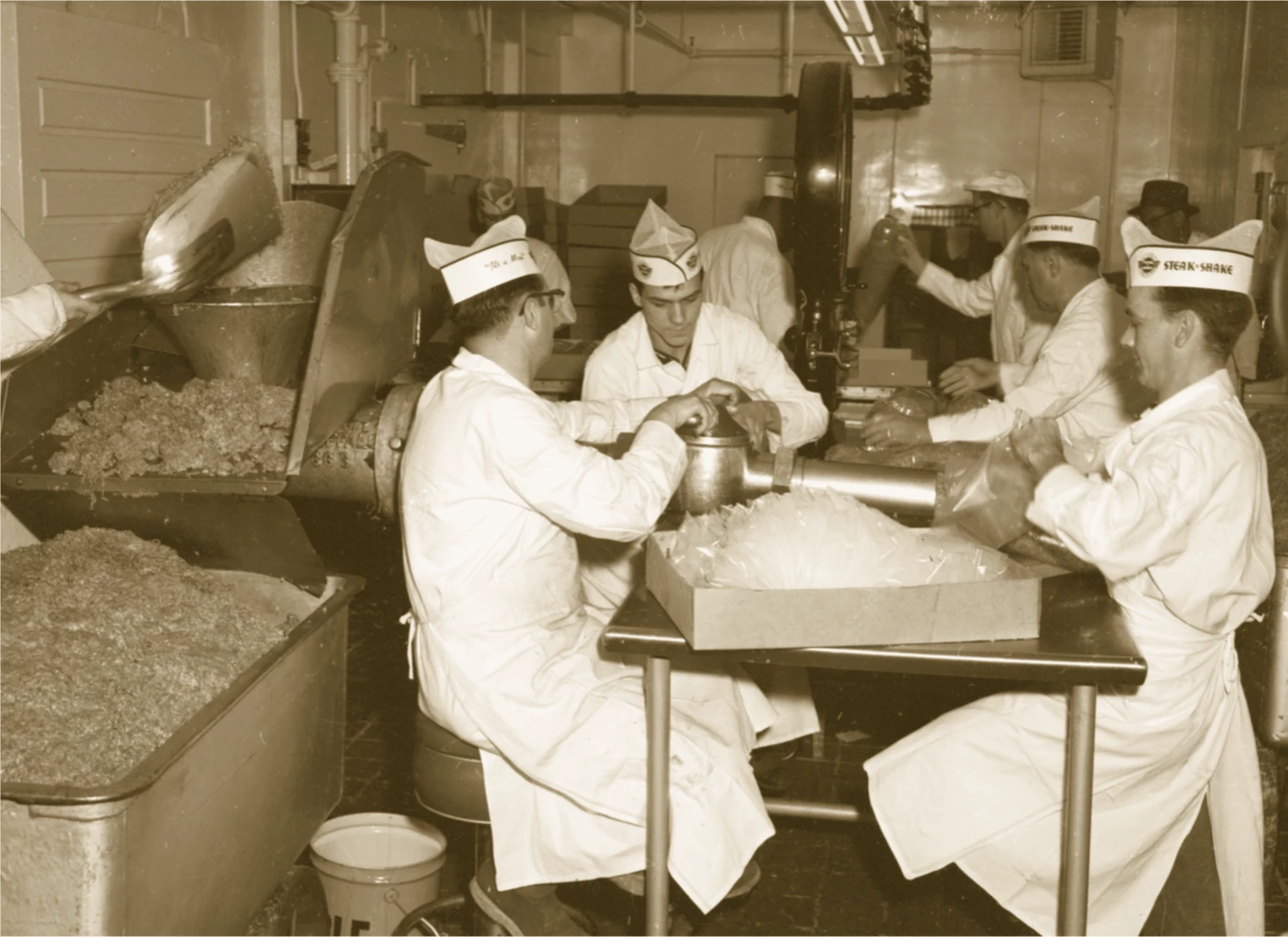 7 people are in a room with industrial equipment and stainless steel tables. In the foreground there are 4 light-skinned men wearing white clothing and white aprons and it appears they are extruding meat into plastic bags. They are all wearing paper hats with the Steak n Shake logo and name. On the left side of the image are two huge containers filled with ground beef. You can see a shovel actively pouring meat from one container to the next.