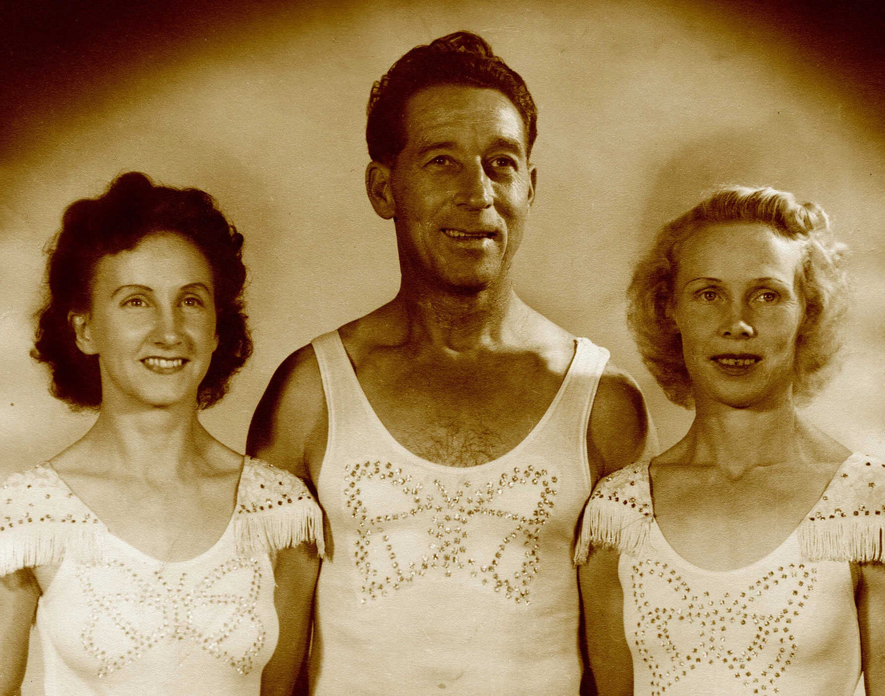Portrait of two women on either side of one man. The woman on the left has short dark hair, the man has short dark hair, and the woman on the right has short light hair. They are all smiling and looking to the right of the camera. They are wearing matching outifts, white skin tight fabric with a butterfly shape in sequins on the chest. The ladies have a fringe detail on the shoulder.