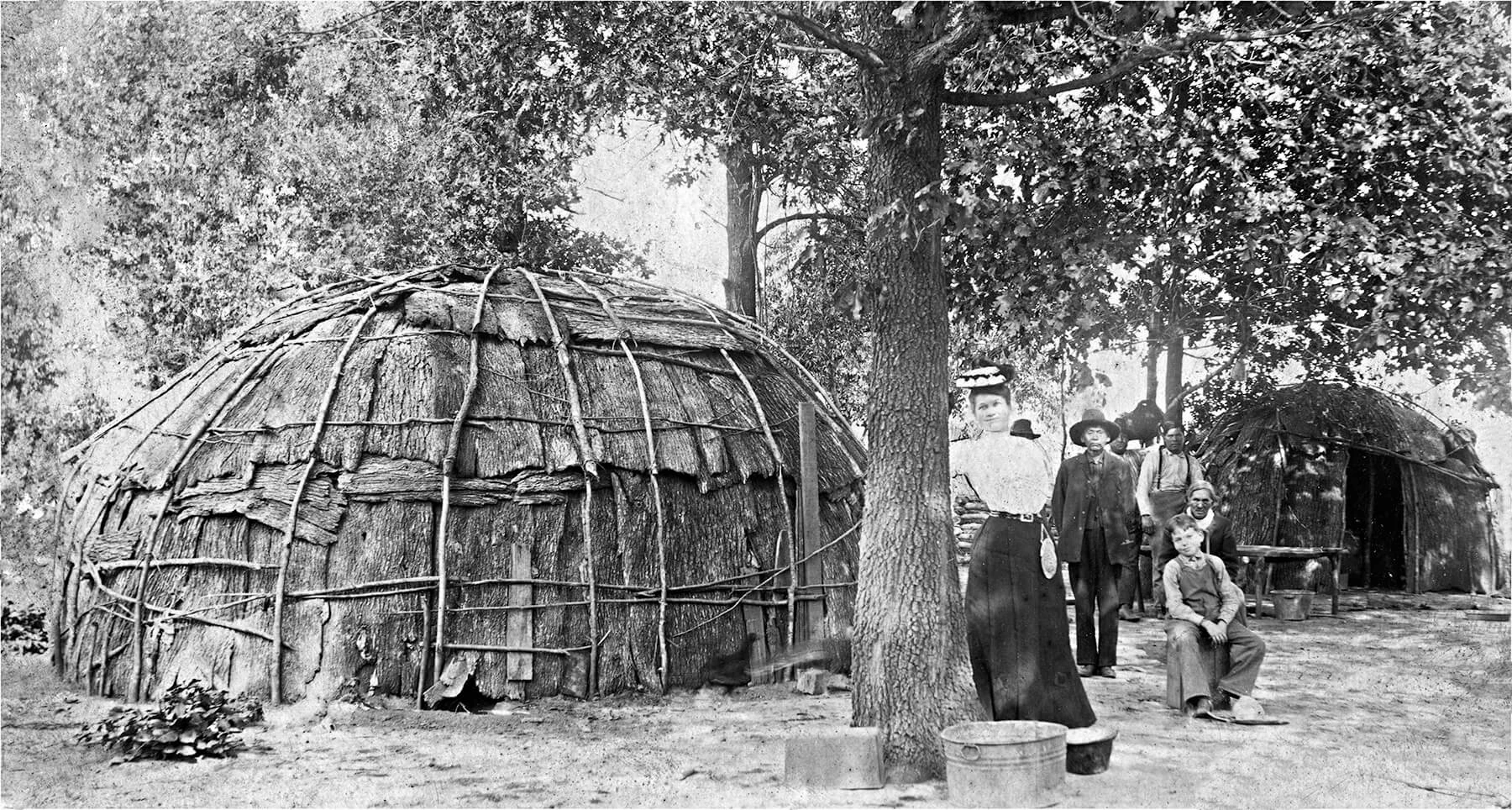 Black and white photo of a group of men and women standing beside a wigwam.